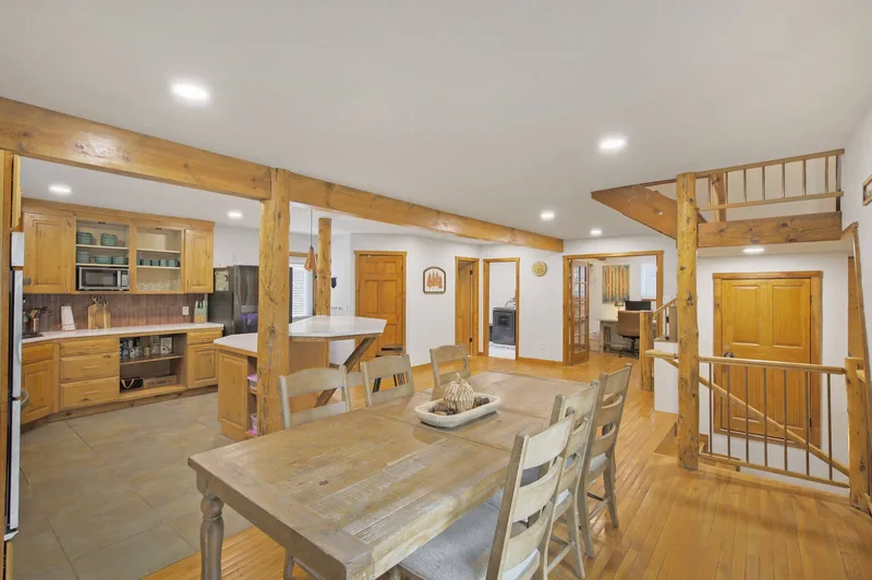 Open dining area beside the kitchen: a long wood table with upholstered chairs on wood floors under an exposed beam, with a sliding door to the deck and forest through the glass