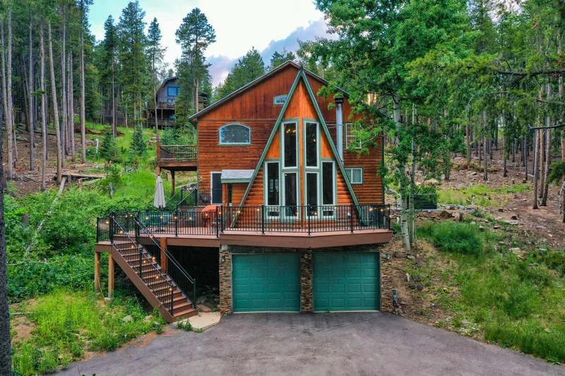 Three-story A-frame clad in orange-brown log siding with teal-green trim and a tall triangular window wall, an elevated deck with black railing, ringed closely by lodgepole pines