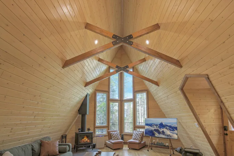 Great room under the A-frame gable: knotty-pine tongue-and-groove walls and vaulted ceiling with exposed cross beams and a ceiling fan, a full-height triangular window wall onto green forest, a black wood-burning stove on a hearth, and a gray sectional sofa on wood floors