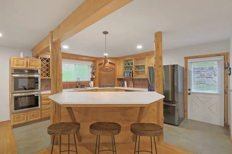 Kitchen with honey-toned wood cabinetry, white solid-surface counters, a center island with breakfast-bar stools, a stainless refrigerator and wall oven, a built-in wine rack, wood posts and beams, and a window over the sink onto forest
