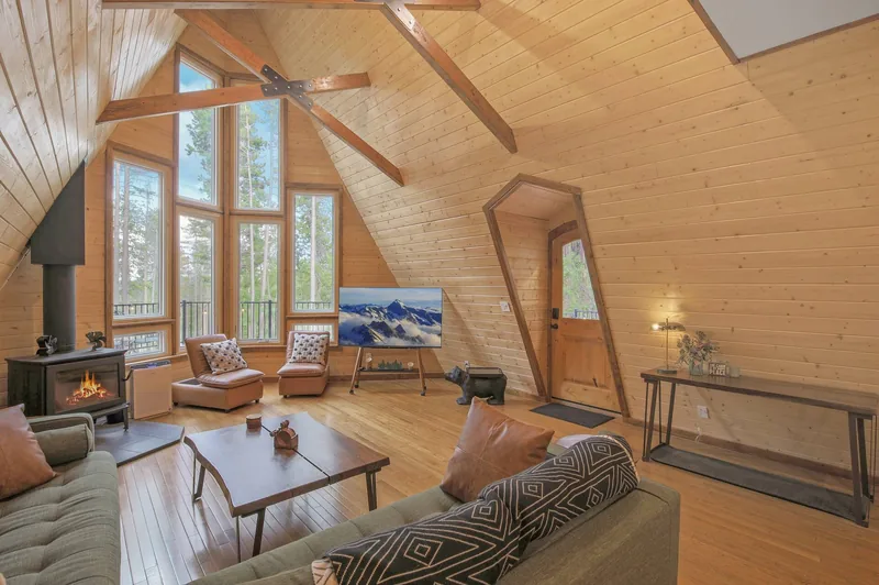 Second living area with a knotty-pine vaulted ceiling and exposed beams, a stone-faced fireplace, an upholstered sofa and armchairs around a wood coffee table, and tall windows onto the forest