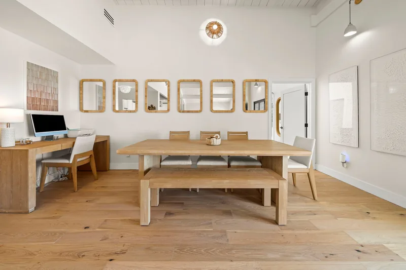 Long light-oak dining table with a bench on one side and woven chairs on the other, under a white vaulted ceiling beside a gallery wall of framed art, on oak floors