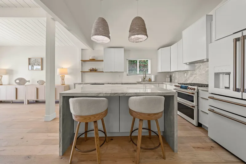 Kitchen with white cabinetry, a marble-look waterfall island with three round-backed wood stools, dome pendant lights, a stainless range and hood, and open oak shelving