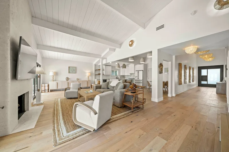Living room with a white vaulted ceiling and exposed white beams, pale wide-plank oak floors, a white plaster fireplace, cream sofas and armchairs, a wall-mounted television, and glazed doors to the outside