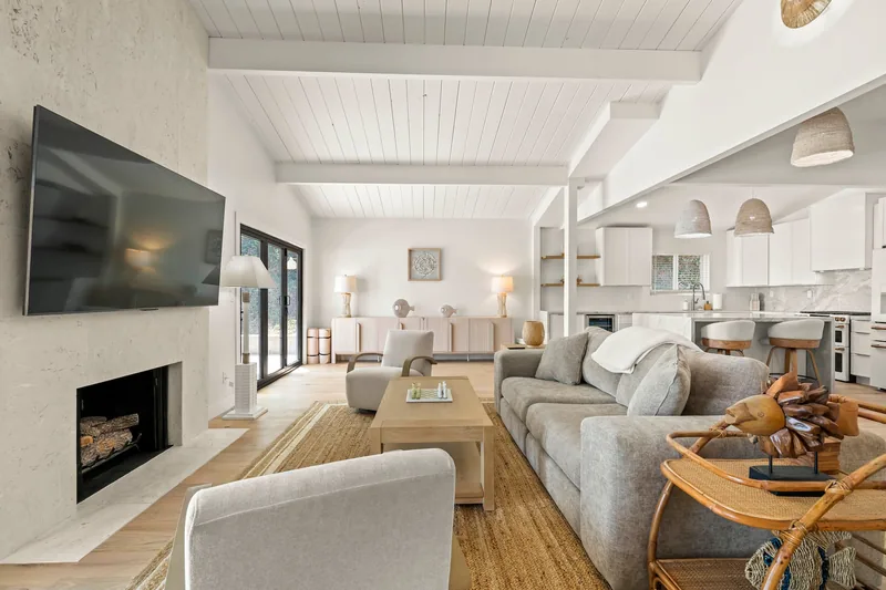 Second view of the living space: white vaulted ceiling, plaster fireplace with the television above it, curved cream armchairs on oak floors, and an open sightline through to the kitchen
