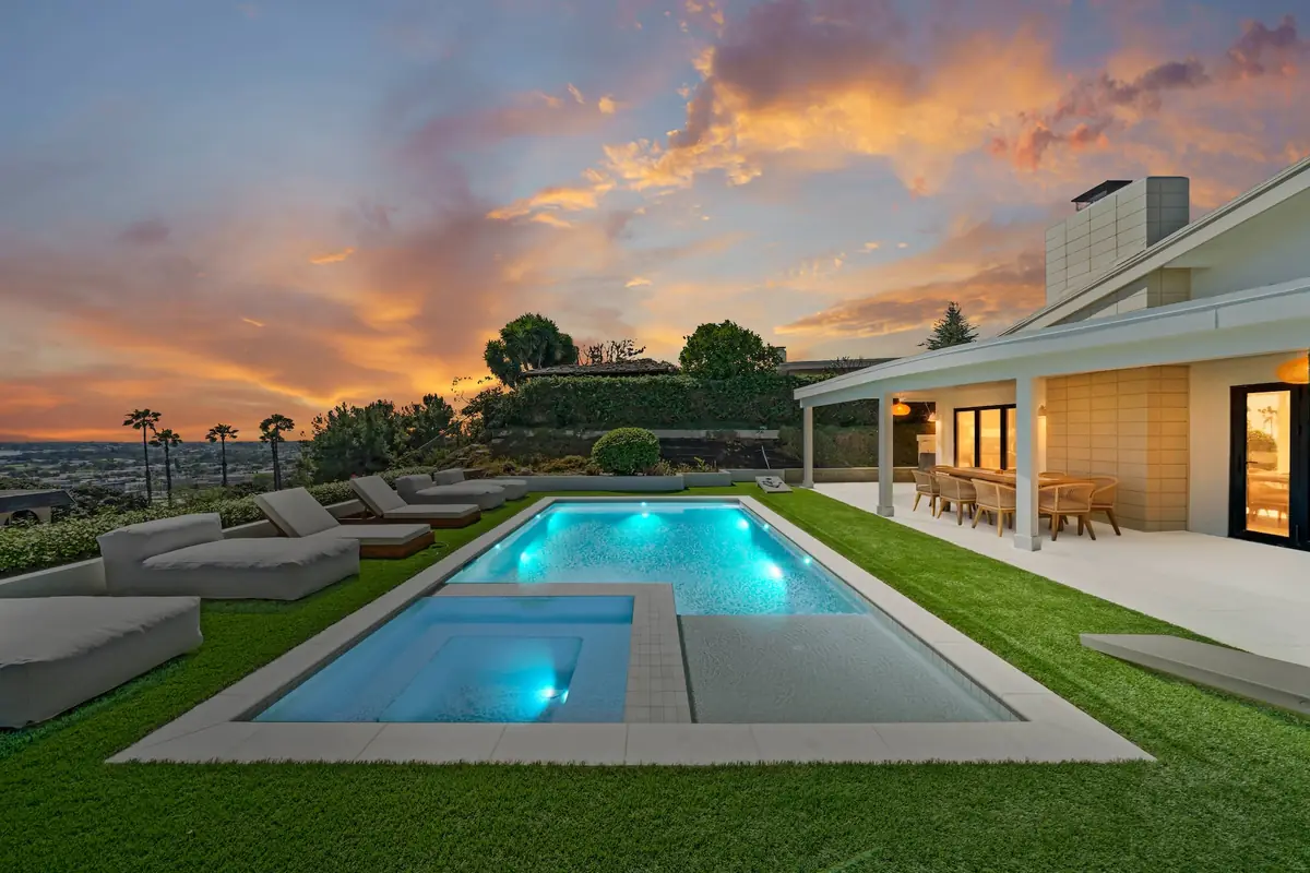 Long rectangular pool lit turquoise at dusk with a square spa at the near end, a single-story white house behind with light spilling from a wall of glass, under an orange and violet sunset sky